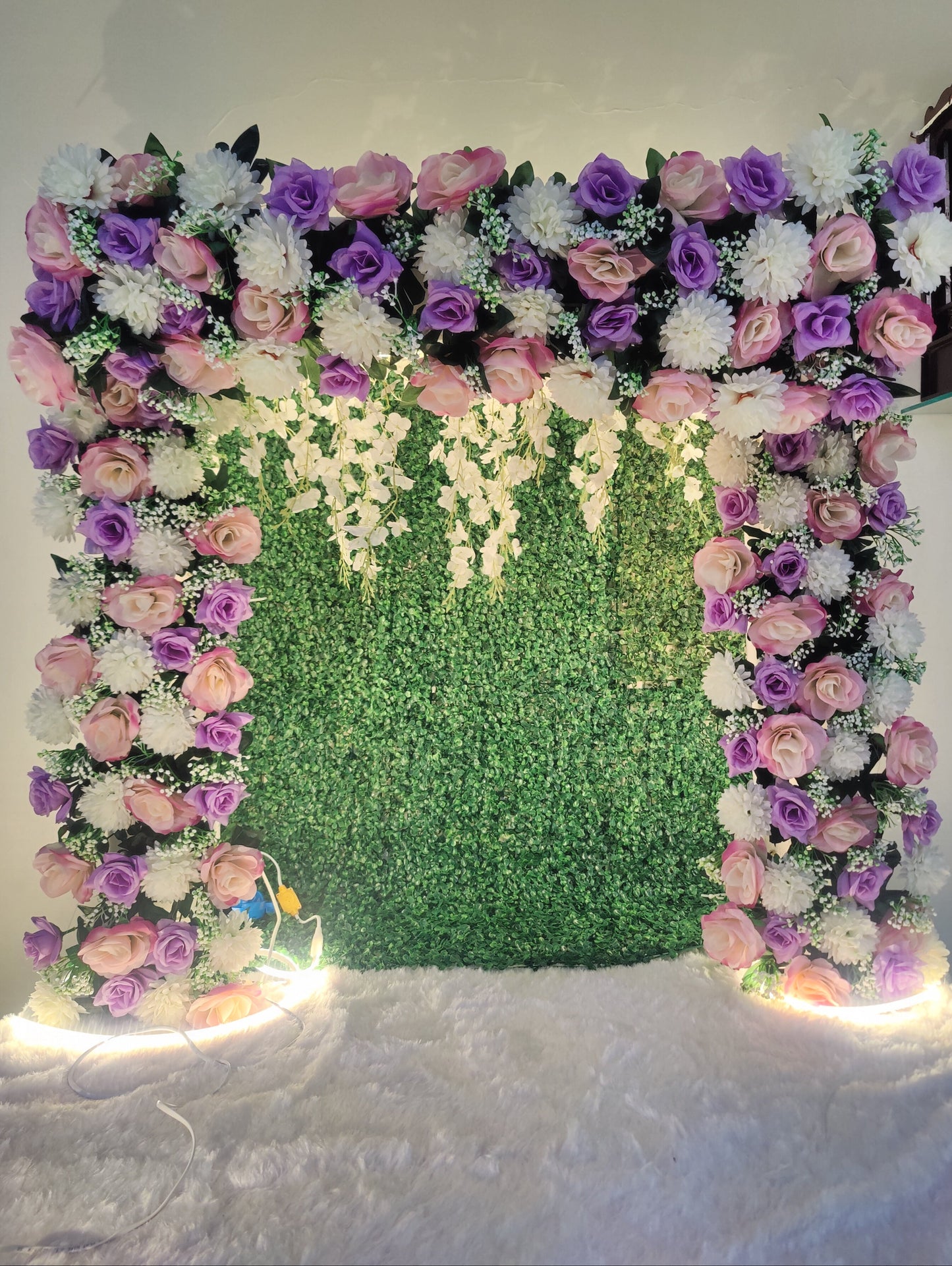 A decorative floral photo backdrop with a green faux grass center, pink and purple roses, white flowers, and warm underlight on a white surface against a plain indoor wall.